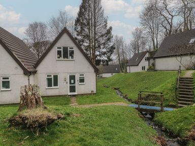 Houses with pitched roofs near a small stream with a wooden bridge and steps in a grassy area at Valley Lodge 38 in St Ann's Chapel Cornwall