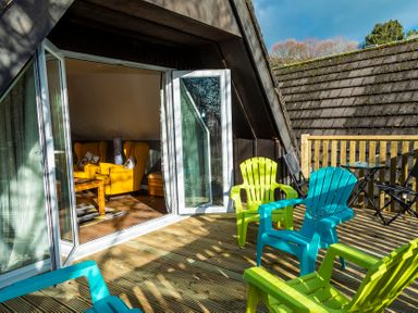 A wooden deck with green and blue plastic chairs outside a room with yellow armchairs at Valley Lodge 40 in St Ann's Chapel Cornwall