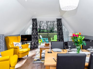 A dining area and living room with yellow chairs a wooden table and glass doors opening to a balcony with plastic chairs at Valley Lodge 40 in St Ann's Chapel Cornwall