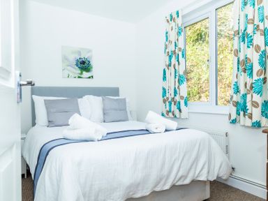 A bedroom with a bed with white and gray pillows and rolled towels on top near a window with floral curtains at Valley Lodge 40 in St Anns Chapel Cornwall