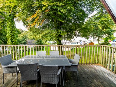 An outdoor wooden deck with a glass-topped table and six wicker chairs surrounded by trees at Valley Lodge 3 St Ann's Chapel Cornwall