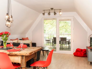 A dining area with a wooden table and red chairs next to a living space with a couch and a small table by glass doors leading to a balcony at Valley Lodge 3 in St Ann's Chapel Cornwall