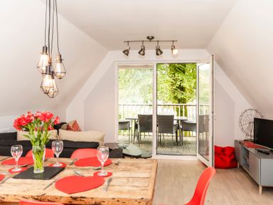 A dining and living room with a wooden table set with red heart-shaped placemats and wine glasses a black sofa and a tv unit with a television and open doors leading to a balcony with outdoor chairs at Valley Lodge 3 in St Ann's Chapel Cornwall