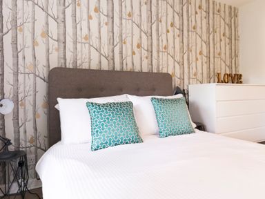 A bedroom with a bed with grey headboard two teal pillows white dresser with love decoration and tree patterned wallpaper at Valley Lodge 3 in St Ann's Chapel Cornwall