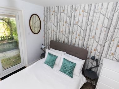 A bedroom with a double bed with white bedding and teal pillows beside a window and a wall with tree patterned wallpaper at Valley Lodge 3 St Ann's Chapel Cornwall