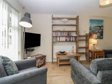 A living room with gray sofas a television mounted on the wall wooden shelves with books and decorations a wooden coffee table and a side table with a lamp at 26 Manorcombe St Ann's Chapel Cornwall