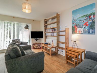 A living room with two grey sofas a wooden bookshelf a wall-mounted television and a painting of a seaside village at 26 Manorcombe in St Ann's Chapel Cornwall