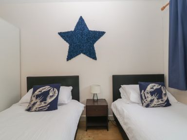 A bedroom with two single beds with white bedding and blue lobster pillows a nightstand with a lamp between the beds and a blue star wall decoration at 26 Manorcombe in St Ann's Chapel Cornwall