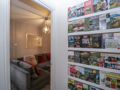 A wall with multiple brochures next to a doorway leading to a living room with a grey sectional sofa and a hanging light at Colliers Cottage in Cinderford