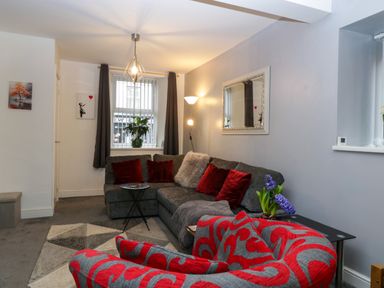 A living room with a grey sectional sofa with red and grey cushions a red patterned armchair a geometric rug and a window with dark curtains at Colliers Cottage in Cinderford