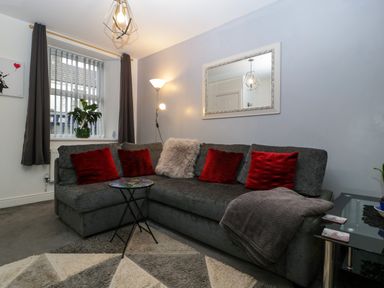 A living room with a gray sectional sofa red pillows a floor lamp a decorative mirror and a small round table at Colliers Cottage in Cinderford