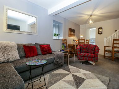 A living room with a gray sectional sofa with red pillows a red patterned armchair a small round table a geometric patterned rug wooden chairs and a dining table with a lamp and wall clock at Colliers Cottage in Cinderford