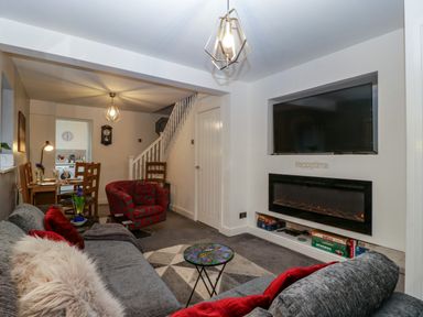 A living room with a gray sofa, a red patterned armchair, a wall-mounted TV, an electric fireplace, a small round table, and a dining area with wooden chairs at Colliers Cottage in Cinderford