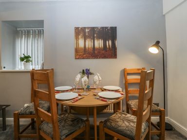 A dining area with a round wooden table set for four with plates and napkins four wooden chairs with patterned cushions and a floor lamp at Colliers Cottage in Cinderford