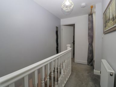 A hallway with a white staircase railing a gray carpet a radiator a door open to another room a curtain beside stairs and a wall painting at Colliers Cottage in Cinderford