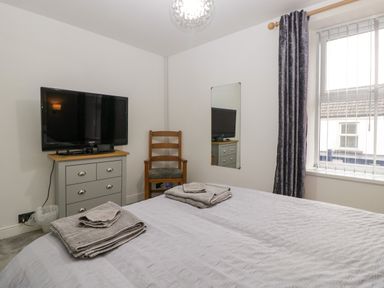 A bedroom with a bed, chest of drawers with a television, wooden chair, window with vertical blinds and curtains at Colliers Cottage in Cinderford