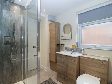 A bathroom with a shower enclosure wooden cabinets a sink a round mirror and a window at Colliers Cottage in Cinderford