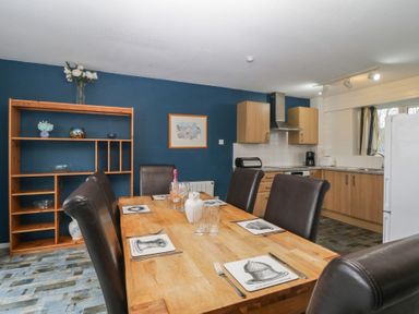 A dining area with a wooden table set with placemats and glasses next to an open kitchen with wooden cabinets at Tamar Cottages 34 in St Ann's Chapel Cornwall
