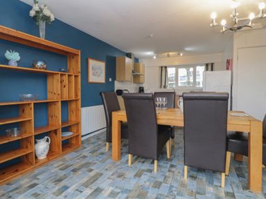 A dining area with a wooden table and black chairs next to a wooden shelving unit and a kitchen with cabinets and a window at Tamar Cottages 34 in St Ann's Chapel Cornwall
