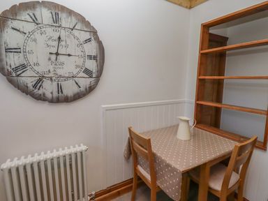 A small dining area with a wooden table covered by a polka dot tablecloth two wooden chairs a white pitcher a large wall clock and a white radiator at Seafield Granary in Alnmouth