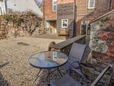 An outdoor patio with gravel ground a round glass table two chairs a stone and brick wall and a wooden bench at Seafield Granary in Alnmouth