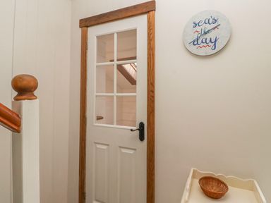 A hallway with a white door with glass panes and wooden trim and a wall clock with text above a small table with a wicker bowl at Seafield Coach House in Alnmouth