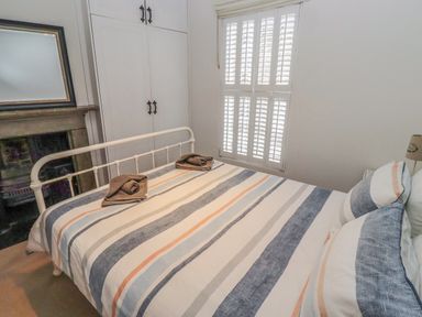 A bedroom with a bed with striped bedding two folded towels on the bed a window with shutters a wardrobe and a fireplace at Seafield Coach House in Alnmouth