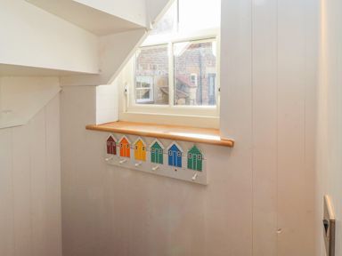A white corner with a window above a wooden ledge and a wall-mounted coat rack with colorful house-shaped hooks at Seafield Coach House in Alnmouth