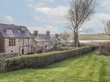 A row of stone houses with a grassy garden and a leafless tree overlooking a river with a boat at Seafield Coach House in Alnmouth