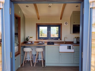 A kitchenette with two stools plates mugs and a sink under a window at Fir Tree Shepherd's Hut in Llanon