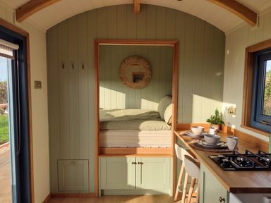 A small kitchen area with stove and counter set with bowls and mugs beside a window and a raised bed with green bedding at Fir Tree Shepherd's Hut in Llanon