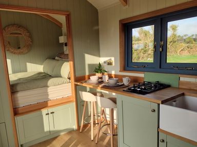 A small kitchen area with a gas stove, sink, two stools at a wooden counter, and a bed with cushions in an alcove at Fir Tree Shepherd's Hut in Llanon