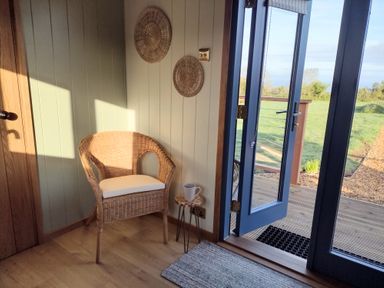 A wicker chair with a white cushion next to a small wooden table with a mug by a blue door opening to a grassy area at Fir Tree Shepherd's Hut in Llanon