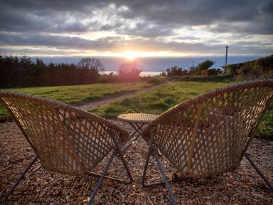 Two wicker chairs and a small table on wood chips facing a grassy field and trees at sunset at Fir Tree Shepherd's Hut in Llanon