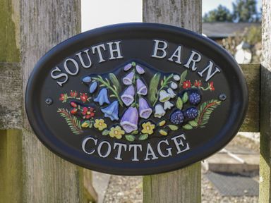 A black oval sign with flowers and berries reading South Barn Cottage on a wooden fence at South Barn Cottage in Hatford near Stanford In The Vale