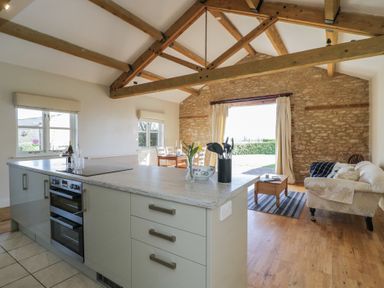 An open kitchen and living area with wooden beams a stone wall a sofa and dining table at South Barn Cottage in Hatford near Stanford In The Vale