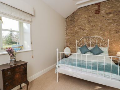 A bedroom with a white metal bed frame against a stone wall and a wooden dresser with flowers on a windowsill at South Barn Cottage in Hatford near Stanford In The Vale