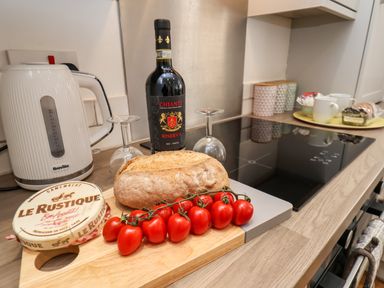 A kitchen countertop with a loaf of bread, cherry tomatoes on a vine, a round box of camembert cheese, a bottle of Chianti wine with two upside-down wine glasses, a white electric kettle, and a yellow tray with mugs and containers at The Bungalow in Whitby