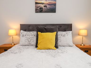 A bedroom with a bed with patterned bedding yellow and gray pillows two bedside tables with lamps and a landscape picture on the wall at The Bungalow in Whitby