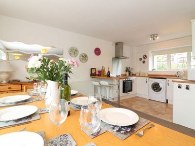 A dining area with a wooden table set with plates and glasses next to an open kitchen with white cabinets and appliances at Tamar Cottages 35 St Ann's Chapel Cornwall