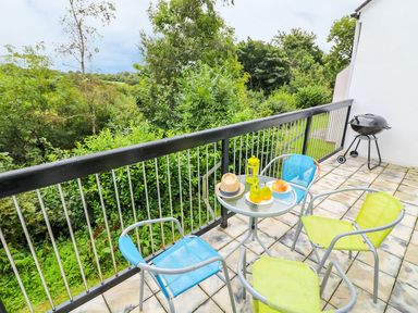 A balcony with a small round glass table with pears croissants and a hat on it three chairs and a barbecue grill with green trees in the background at Tamar Cottages 35 in St Ann's Chapel Cornwall
