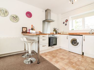 A kitchen with white cabinets a washing machine an oven bar stools and wall decorations at Tamar Cottages 35 in St Ann's Chapel Cornwall