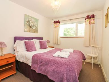 A bedroom with a double bed with purple bedding and pillows a wooden bedside table with a lamp a window with floral curtains and a white chair in the corner at Tamar Cottages 35 St Ann's Chapel Cornwall