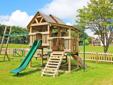 An outdoor wooden playground structure with slides swings and climbing elements in a fenced grassy area at Tamar Cottages 35 in St Ann's Chapel Cornwall