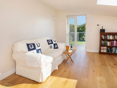 A room with a white sofa with alphabet cushions a small wooden table with drinks and snacks a bookshelf and glass doors leading outside at Moult Hill Barn in Salcombe