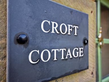A close up of a black metal plaque with Croft Cottage written on it attached to a stone wall near a green window at Croft Cottage in Alnwick