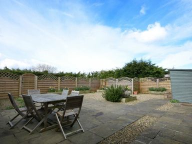An outdoor patio area with a wooden table and six chairs surrounded by a wooden fence and garden beds at Croft Cottage in Alnwick