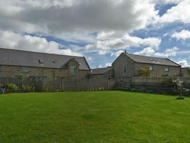 A large green grassy backyard with wooden fences and stone buildings under a partly cloudy sky at Croft Cottage in Alnwick