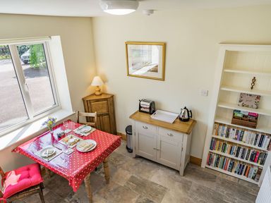 A dining area with a table set for two near a window a cabinet with a toaster and kettle and a bookshelf filled with books at Pant Y Cwtsh in Kidwelly