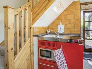 A small kitchen area with red cabinets a sink a microwave and a stove next to wooden stairs and a door to outside at Pant Y Cwtsh in Kidwelly
